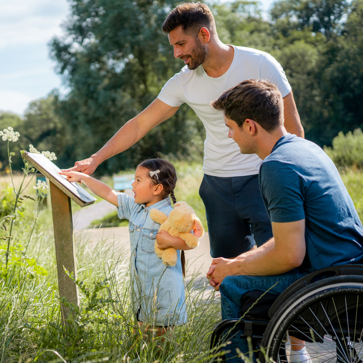 Two men, one standing and one in a wheelchair, look at a sign outside with a little girl